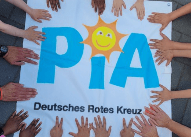 Mehrere Kinder- und Erwachsenenhände legen sich rund um ein Banner mit der Aufschrift „PIA“ und einem lachenden Sonnensymbol. Darunter steht „Deutsches Rotes Kreuz“.