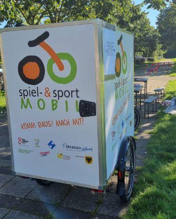 Ein Anhänger mit der Aufschrift „spiel- & sport MOBIL“ steht auf einer Wiese. Die Seiten sind mit einem bunten Logo in Form eines Fahrrads und mehreren Sponsorenlogos bedruckt. Im Hintergrund sind Tische, Bänke und Spielgeräte zu sehen.
